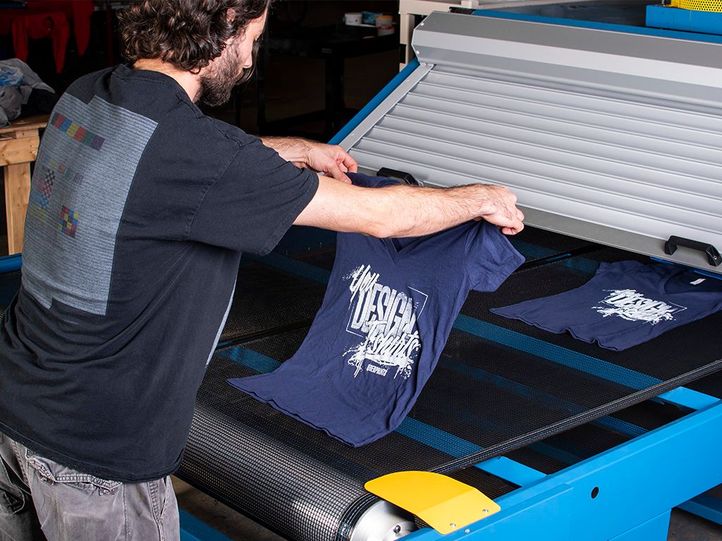 A press operator placing printed t-shirt onto a gas belt dryer.