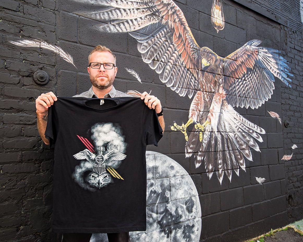 Artist Thomas Turner holding up his t-shirt standing in front of his mural painted for the OuterSpace project.