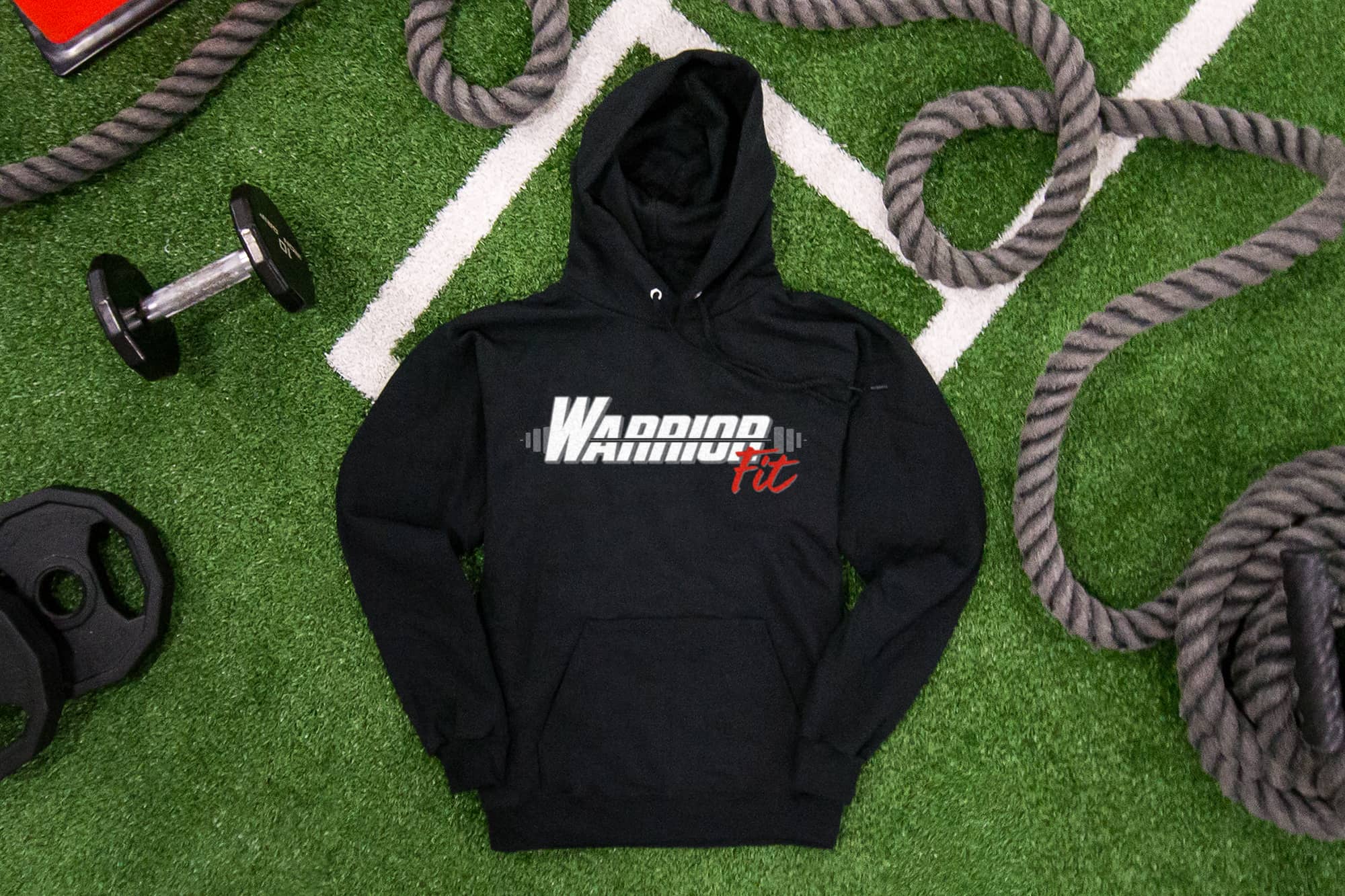 Flatlay of on fake grass of a hoodie with a logo for a gym that reads "Warrior Fit". Weight and other exercise equipment are laying around.