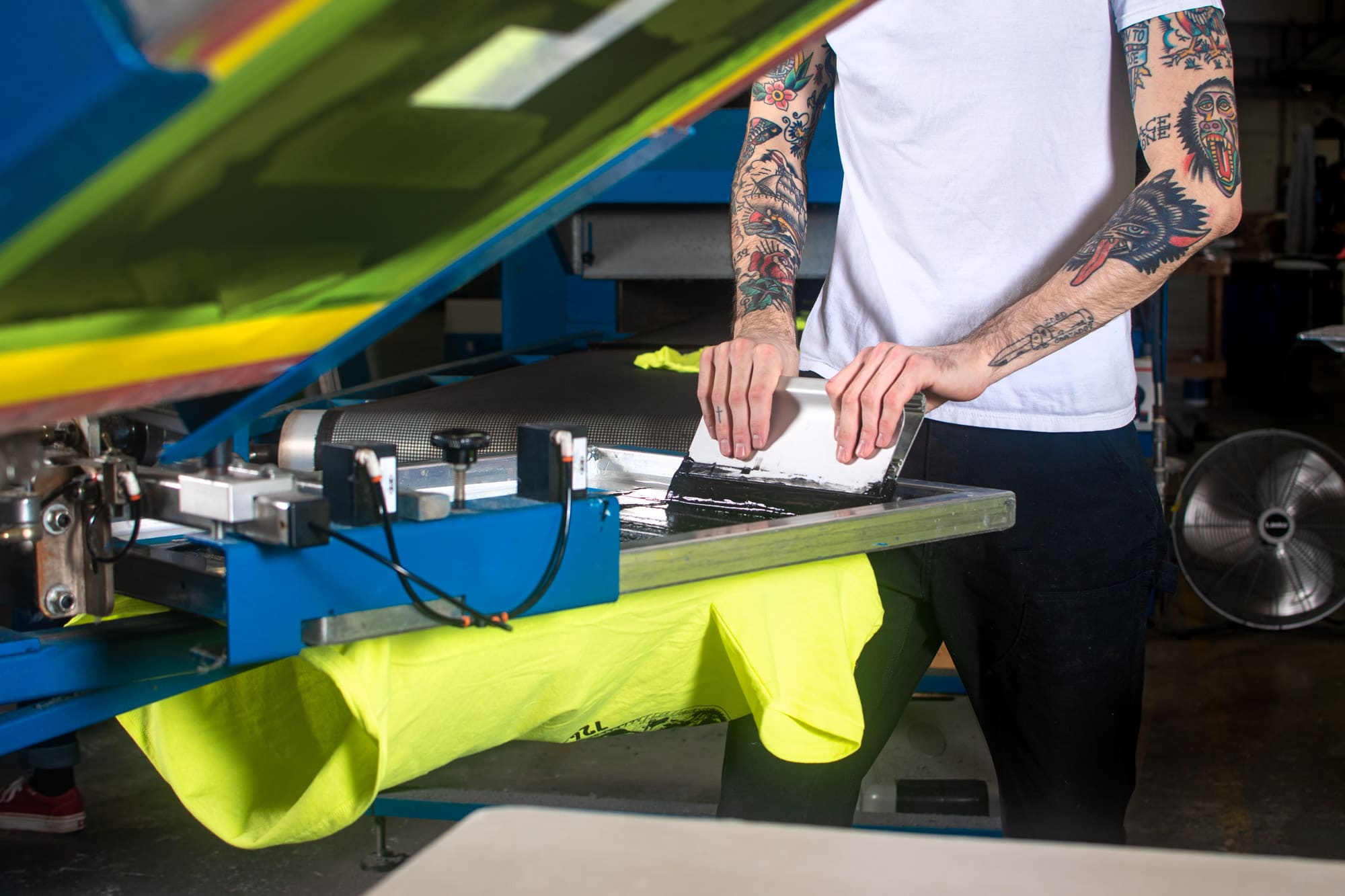 A person with tattoos screen printing a brightly colored t-shirt.