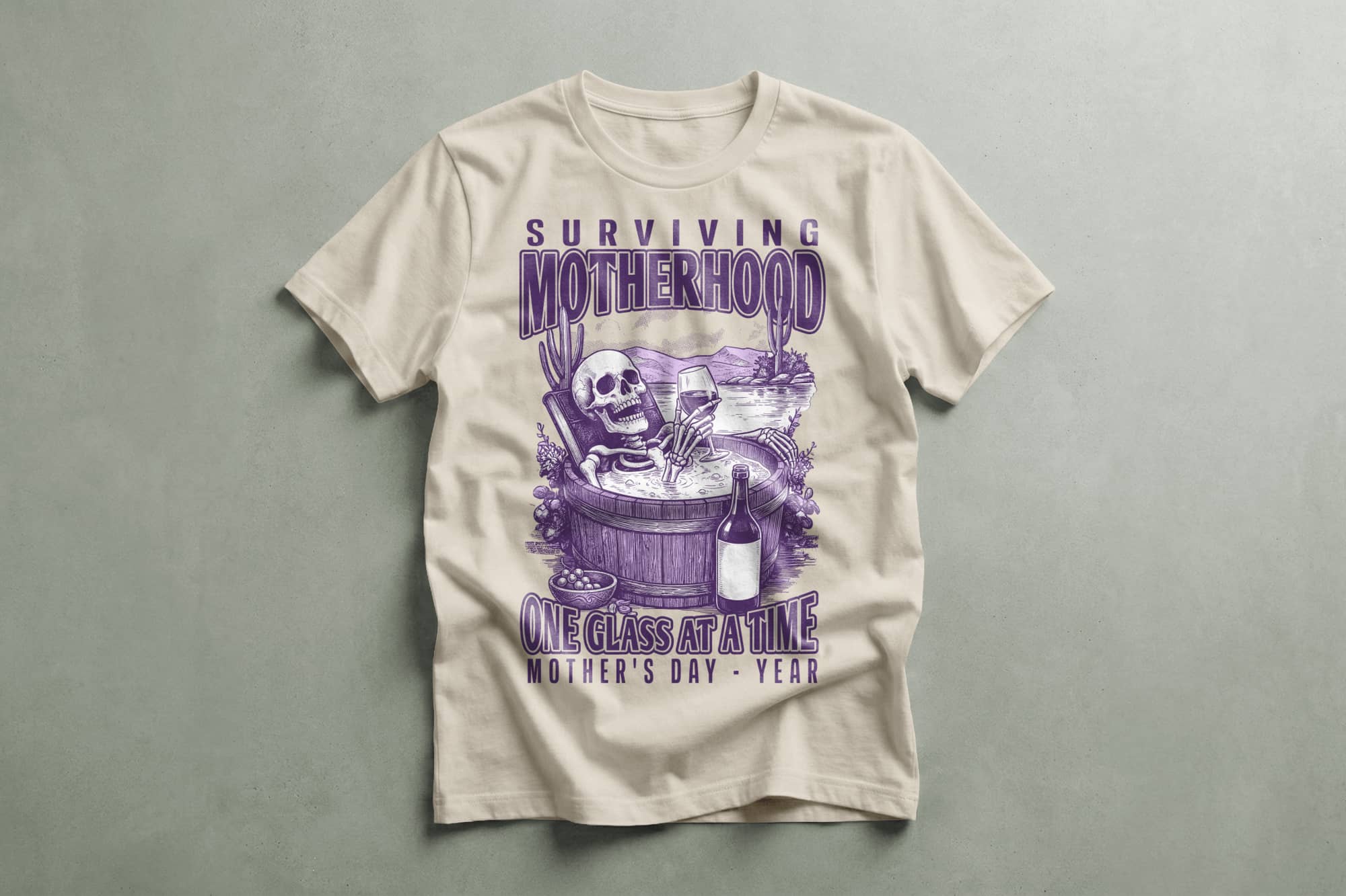 Beige T-shirt with purple graphic of a skeleton in a wine barrel, holding a glass. Text: "Surviving Motherhood One Glass at a Time, Mother's Day." Humorous tone.