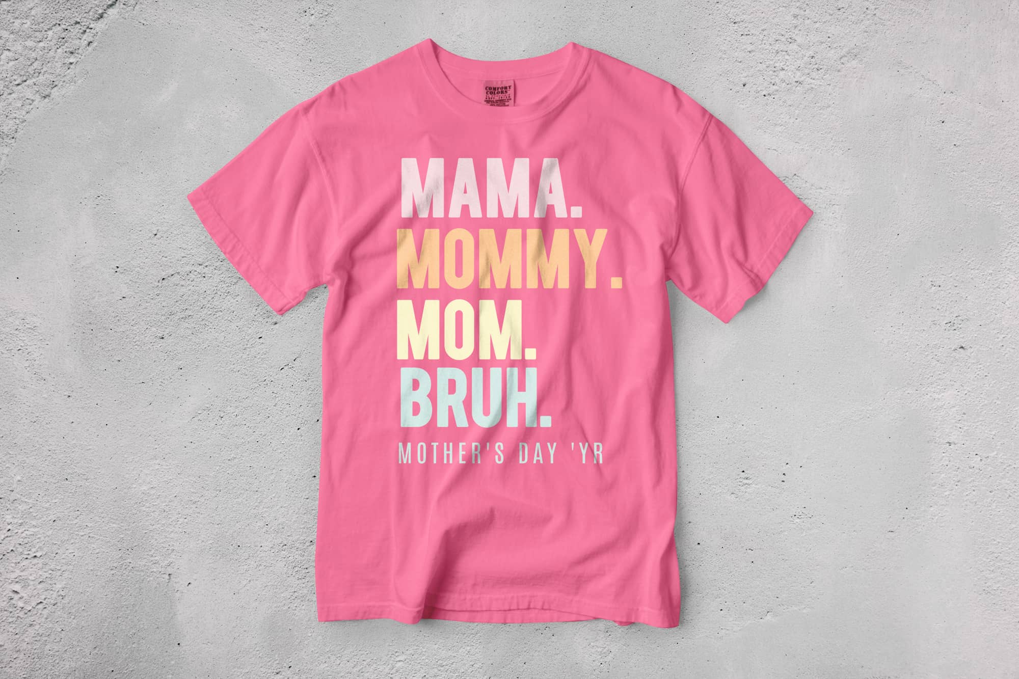 Pink T-shirt with bold text reading, "Mama. Mommy. Mom. Bruh." in white, yellow, and blue on a gray background, with "Mother's Day 'YR" below.
