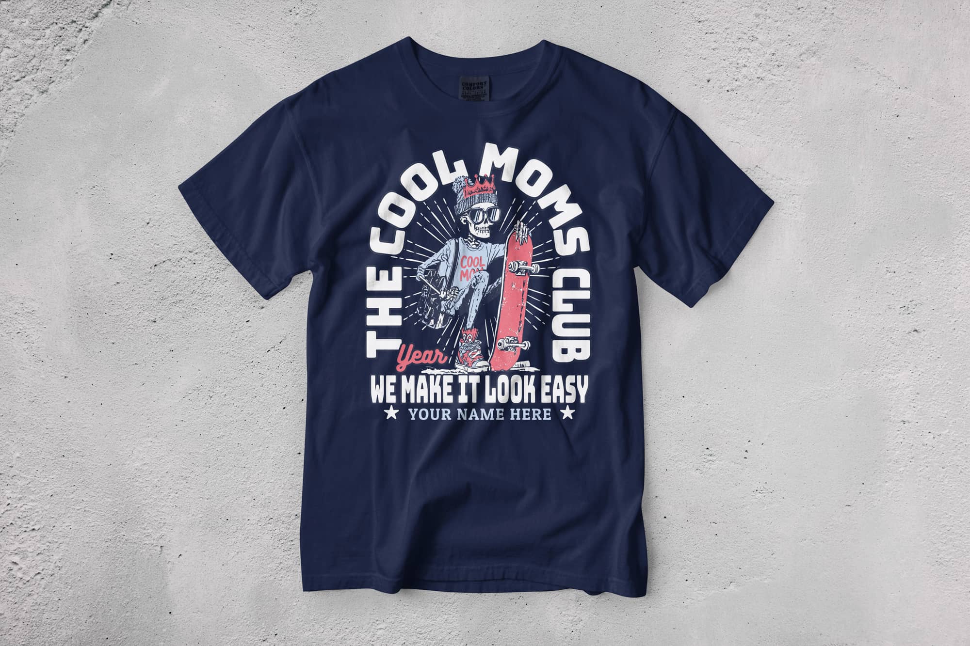 Navy blue T-shirt on a textured surface features a skeleton with a crown, "The Cool Moms Club" text, and a skateboard. Playful and bold design.