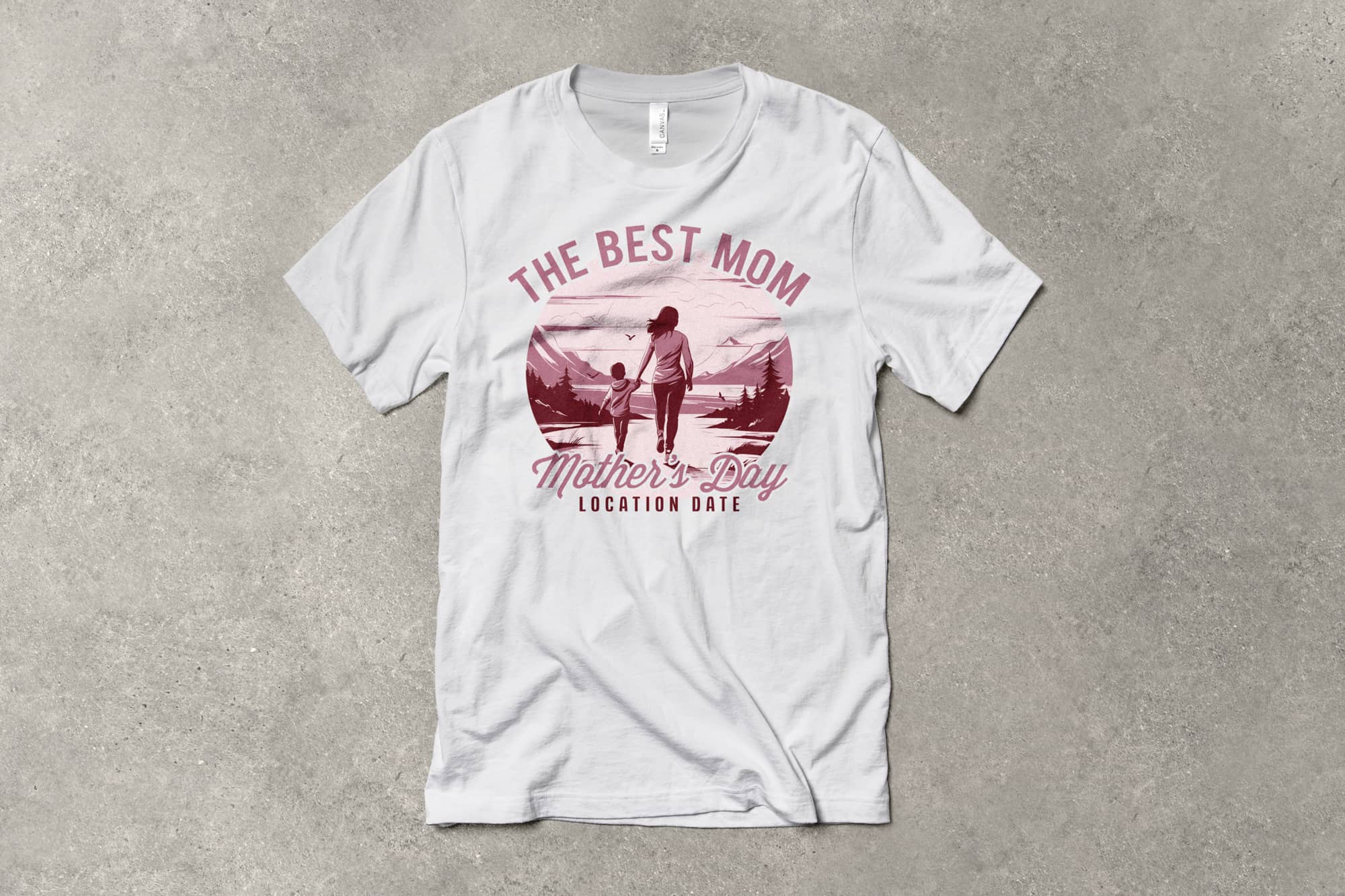 White T-shirt with a pink graphic of a mom and child by a mountain. Text reads "The Best Mom" and "Mother's Day Location Date." Calm, celebratory tone.