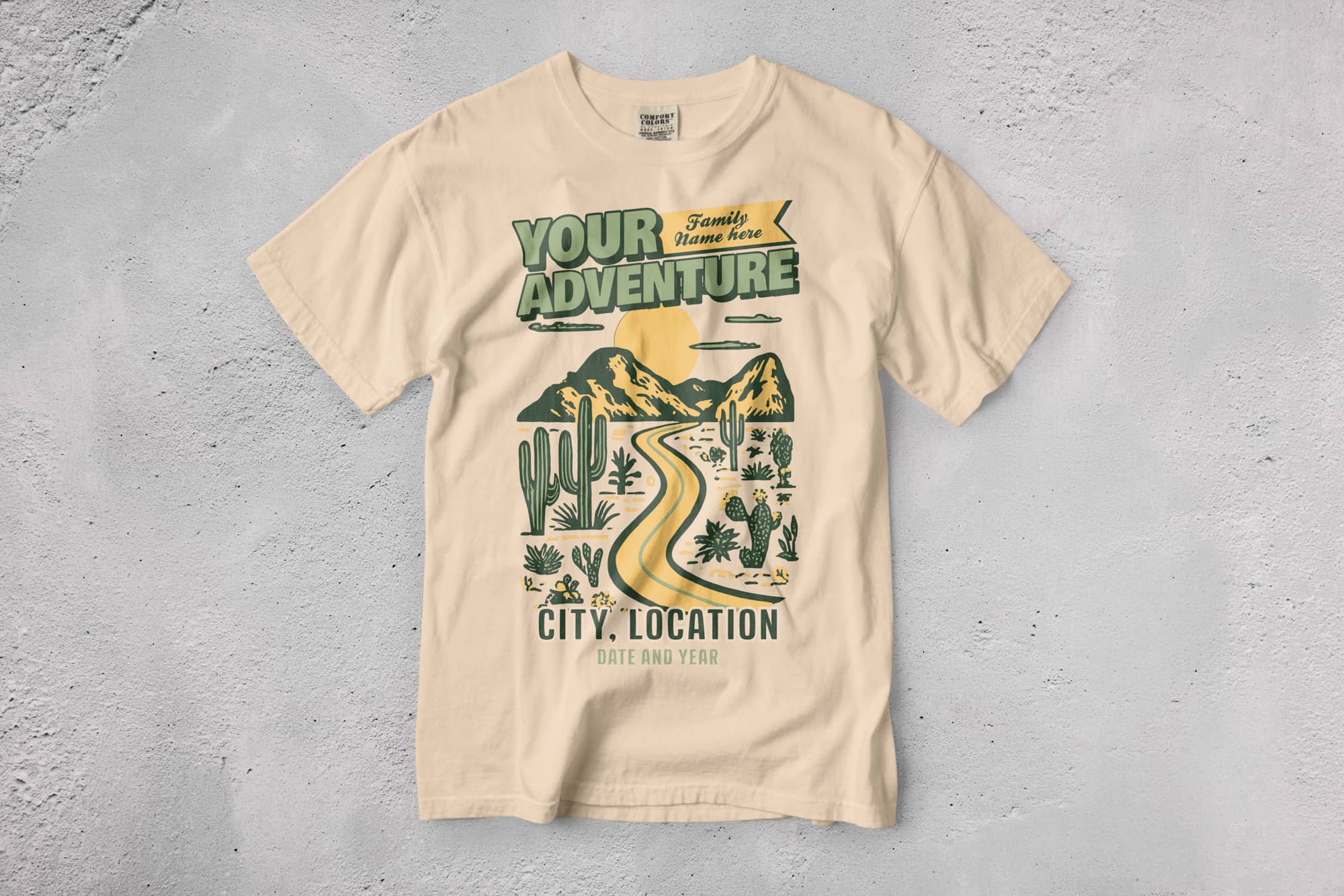 Cream T-shirt on a textured surface featuring a desert scene with cacti, mountains, and a winding road. Bold text reads "Your Adventure," with customizable details below.